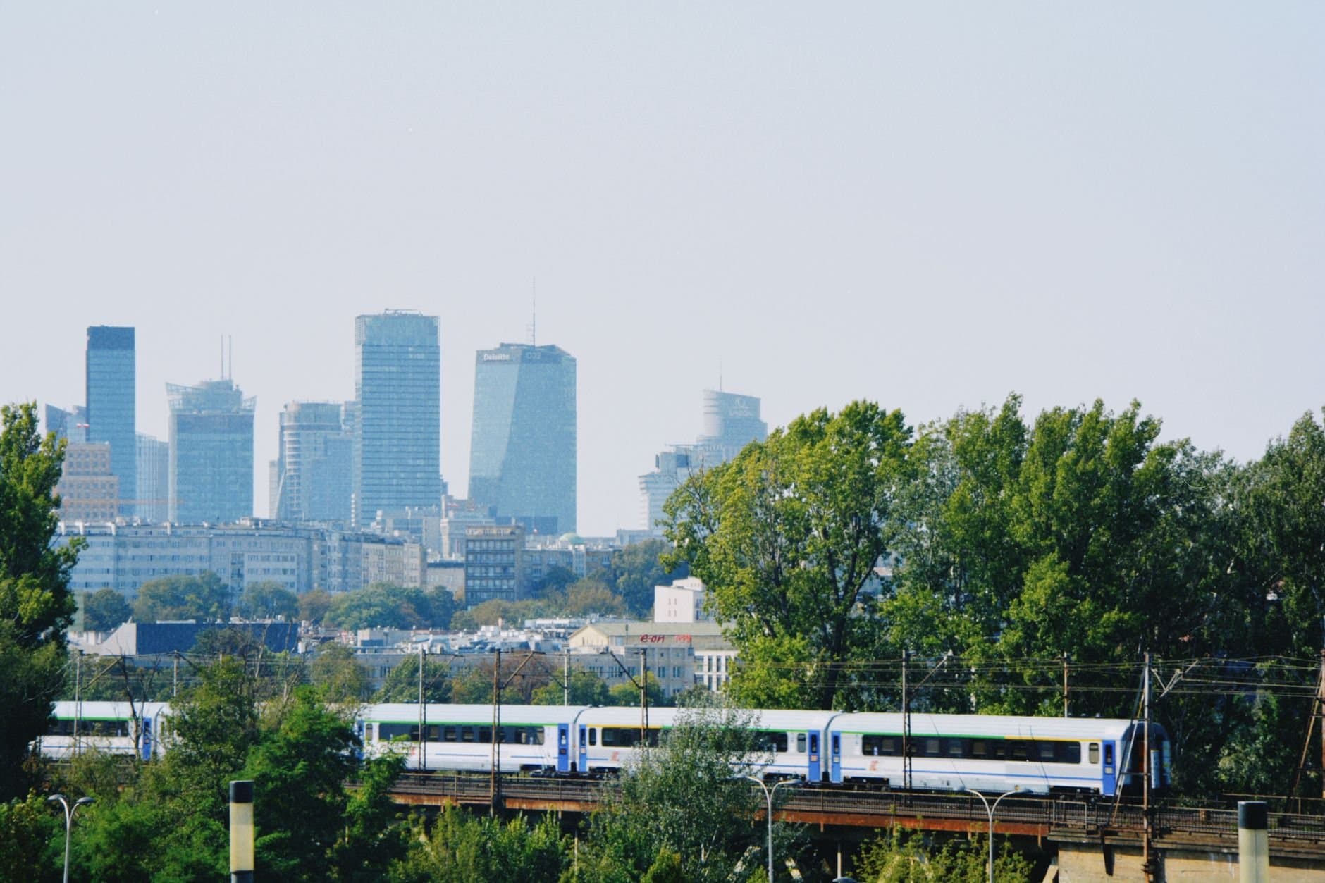 Skyline of Warsaw with modern buildings and a train passing amidst the greenery.